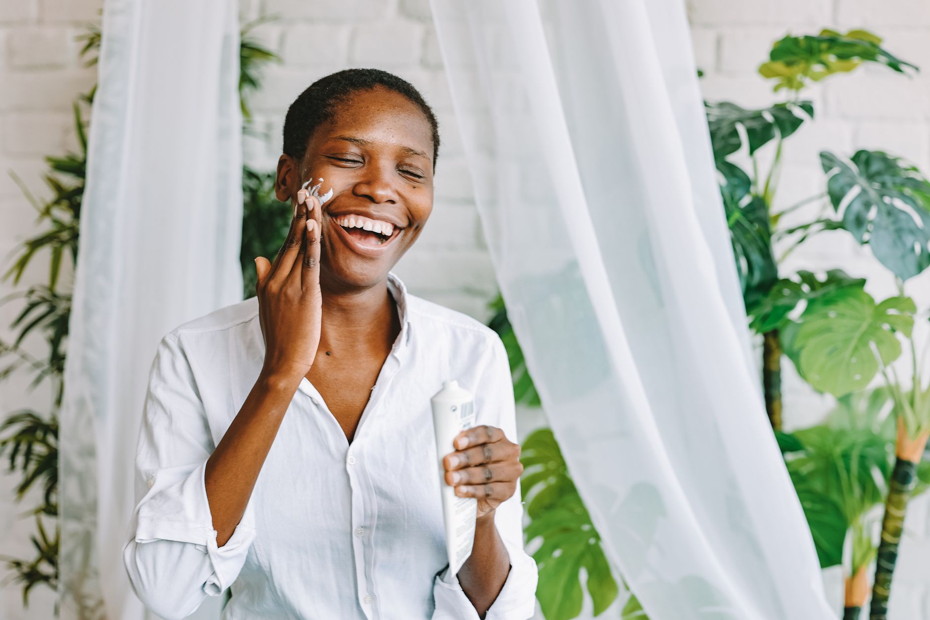 Smiling woman enjoying skincare routine with face cream in a bright, indoor setting with plants.